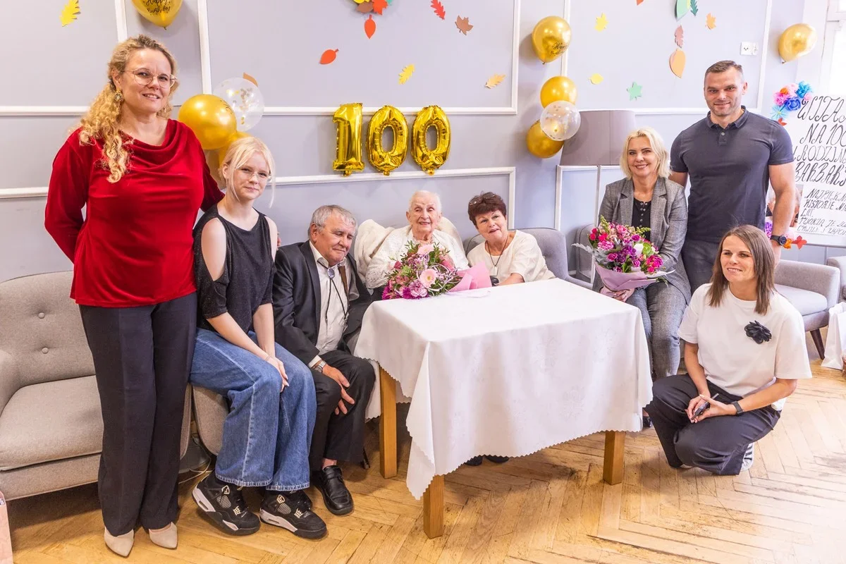 Grupa osób pozuje do zdjęcia podczas uroczystości setnych urodzin w pomieszczeniu ozdobionym balonami i girlandą z cyframi 100.