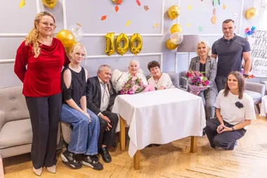 Grupa osób pozuje do zdjęcia podczas uroczystości setnych urodzin w pomieszczeniu ozdobionym balonami i girlandą z cyframi 100.