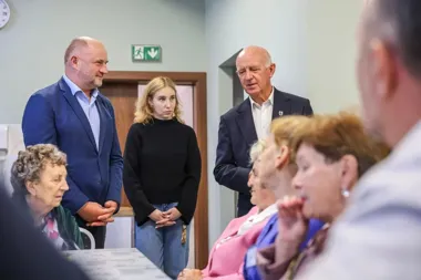 Trzy osoby stoją w pomieszczeniu i rozmawiają z grupą siedzących osób, prawdopodobnie seniorów.